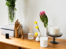 Load image into Gallery viewer, Apotheke Amber Woods votive 2.5oz candle, signature 11oz candle, 3-wick 32oz candle decorating room with stack of books and plants. Wooden hand figure doing OKAY sign and the other doing a sort of peace sign. 3-wick candle is on glass pedestal. Life size comparison.

