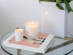 Apotheke Amber Woods votive 2.5oz candle, signature 11oz candle on 2 kinfolk books and on top of glass table top with decorative plant in a tall rectangular white vase.