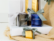 Load image into Gallery viewer, Apotheke Charred Fig 11oz candle and Apotheke Assam Tea 11oz candle in their box packaging each with bowtie on. They sit on top of white platform on top of a sweater material like cloth.
