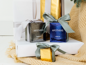 Apotheke Charred Fig 11oz candle and Apotheke Assam Tea 11oz candle in their box packaging each with bowtie on. They sit on top of white platform on top of a sweater material like cloth.