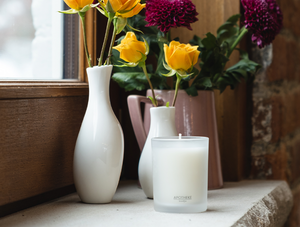 Apotheke Amber Woods signature 11oz candle with decorative flowers in white vases next to it by the window on top of a wooden platform with flowers in grey vase and brick wall in background.