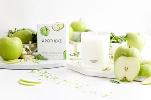 Apotheke Arugula and Green Apple 11oz candle unlit on a white marble platform next to actual green apples. Green apple blurred in the white background. On the left, a bowl of arugula and another green apple sitting on top.