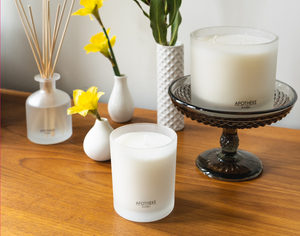 Apotheke signature 11oz candle, 3-wick 32oz candle on wooden table top with decorative flowers and Apotheke diffuser.